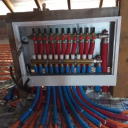 Rozdzielacz ogrzewania podłogowego z czerwono-niebieskimi rurami w izolacji, zamontowany w białej skrzynce, widoczne rury rozprowadzone na siatce w pomieszczeniu ze skośnym dachem.