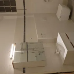 Nowoczesna, biała łazienka z umywalką w zabudowie z płytek, szafką z lustrem i podwieszaną toaletą, wykończona w jasnych odcieniach z akcentami ciemnych płytek w pionowych pasach.