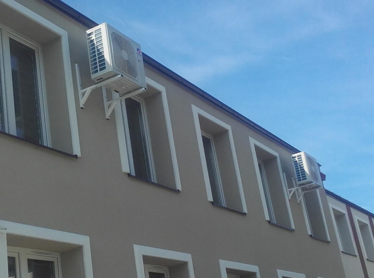 Dwa klimatyzatory marki GREE zamontowane na elewacji budynku z beżową farbą i białymi obramowaniami okien, widoczne białe wsporniki montażowe i błękitne niebo w tle.