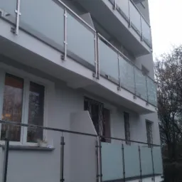 Nowoczesny budynek wielorodzinny z balkonami zabezpieczonymi szklanymi balustradami z metalowymi elementami, widok z dołu na kilka pięter, jesienne drzewo w tle.