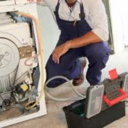 Mężczyzna w kombinezonie roboczym pochylony nad otwartą pralką z widocznym bębnem, skrzynka narzędziowa obok, przewody i węże na podłodze.