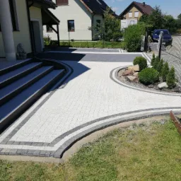 Nowoczesny podjazd z jasnoszarej kostki brukowej, obrzeża z ciemnoszarej kostki, rabata z kamieniami i niskimi krzewami, dom z kolumnami w tle.