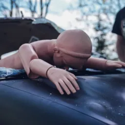 Manekin ratowniczy częściowo wystający przez rozbitą przednią szybę ciemnego samochodu, instruktor w tle.