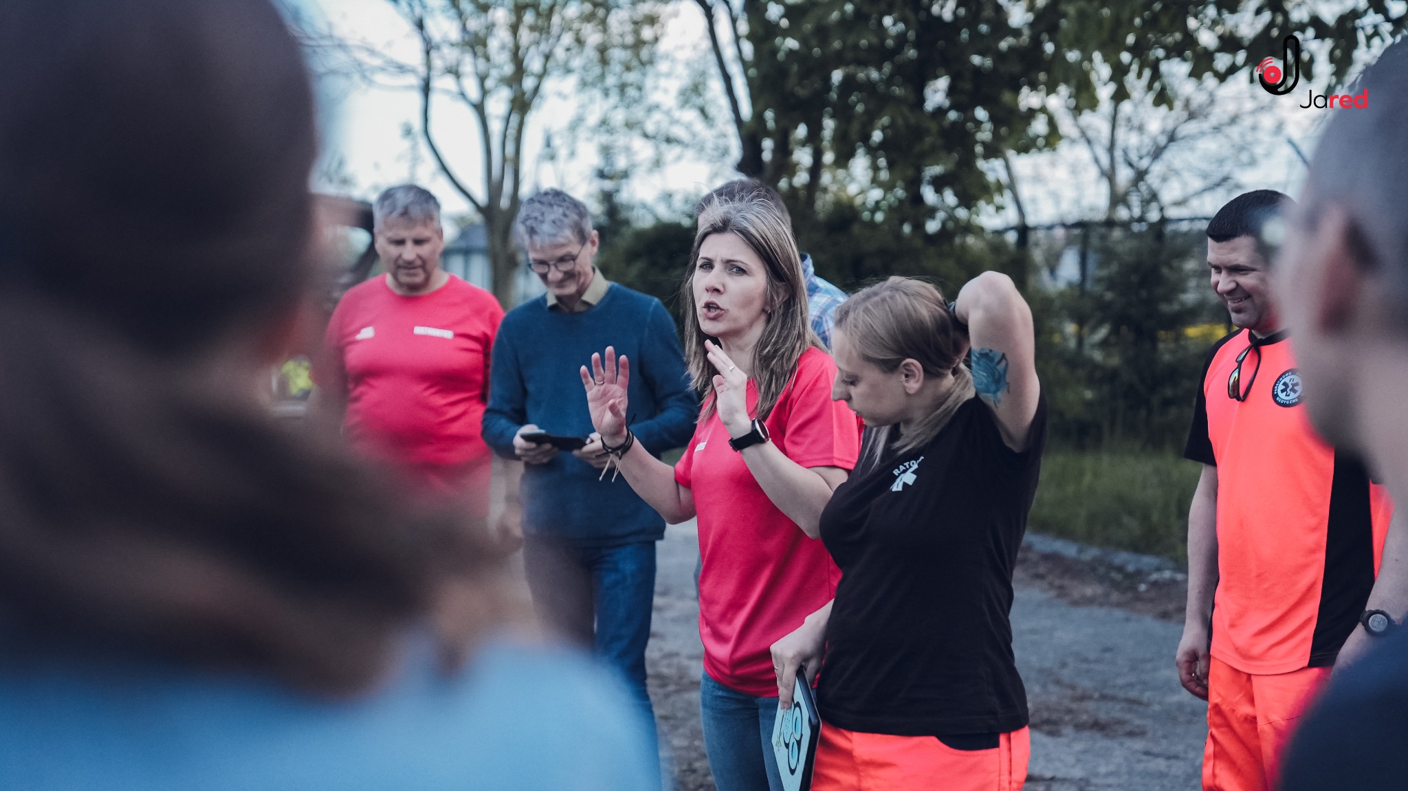 Grupa osób słucha instrukcji instruktorki w różowej koszulce podczas szkolenia na zewnątrz, w tle drzewa i budynki.