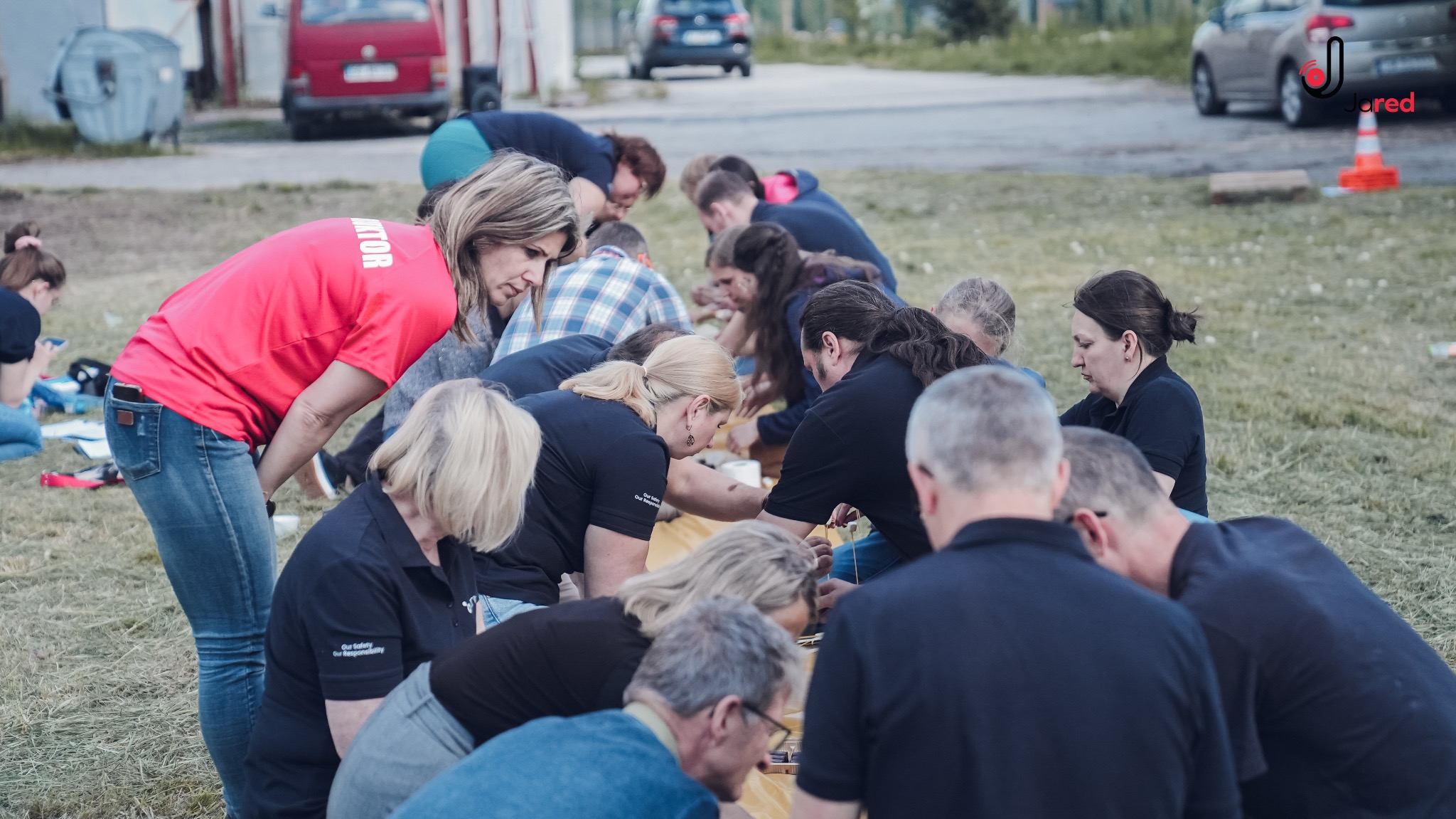 Grupa osób, w tym instruktor w czerwonej koszulce z napisem 'INSTRUKTOR' na plecach, uczestniczy w praktycznym szkoleniu na zewnątrz na trawiastym terenie, skupiona wokół żółtej płachty. W tle...
