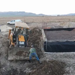 Żółta koparko-ładowarka CAT z operatorem wyrównuje teren pod fundament, widoczny wykop z izolacją, w tle krajobraz polny i biały samochód dostawczy.