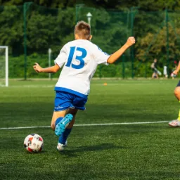 Chłopiec w białej koszulce z numerem 13 i niebieskich spodenkach kopie piłkę na zielonym boisku podczas meczu piłkarskiego, drugi gracz w żółto-zielonym stroju w tle.