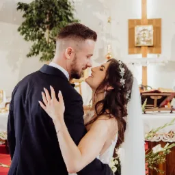 Panna młoda w sukni ślubnej z wiankiem we włosach obejmuje pana młodego w kościele, w tle ołtarz i świece.