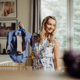 Uśmiechnięta kobieta w kwiecistej sukience trzyma na wieszaku niemowlęce ubranko: błękitne body z długim rękawem, beżowe spodnie i krawat w kratę, w tle łóżeczko dziecięce i okno z widokiem na zieleń.