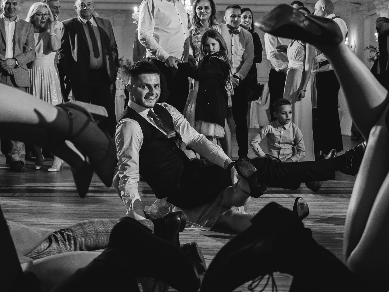 Czarno-białe zdjęcie z wesela, na pierwszym planie uśmiechnięty mężczyzna w kamizelce leżący na podłodze, wokół niego nogi gości w różnych pozach, w tle obserwujący goście weselni.