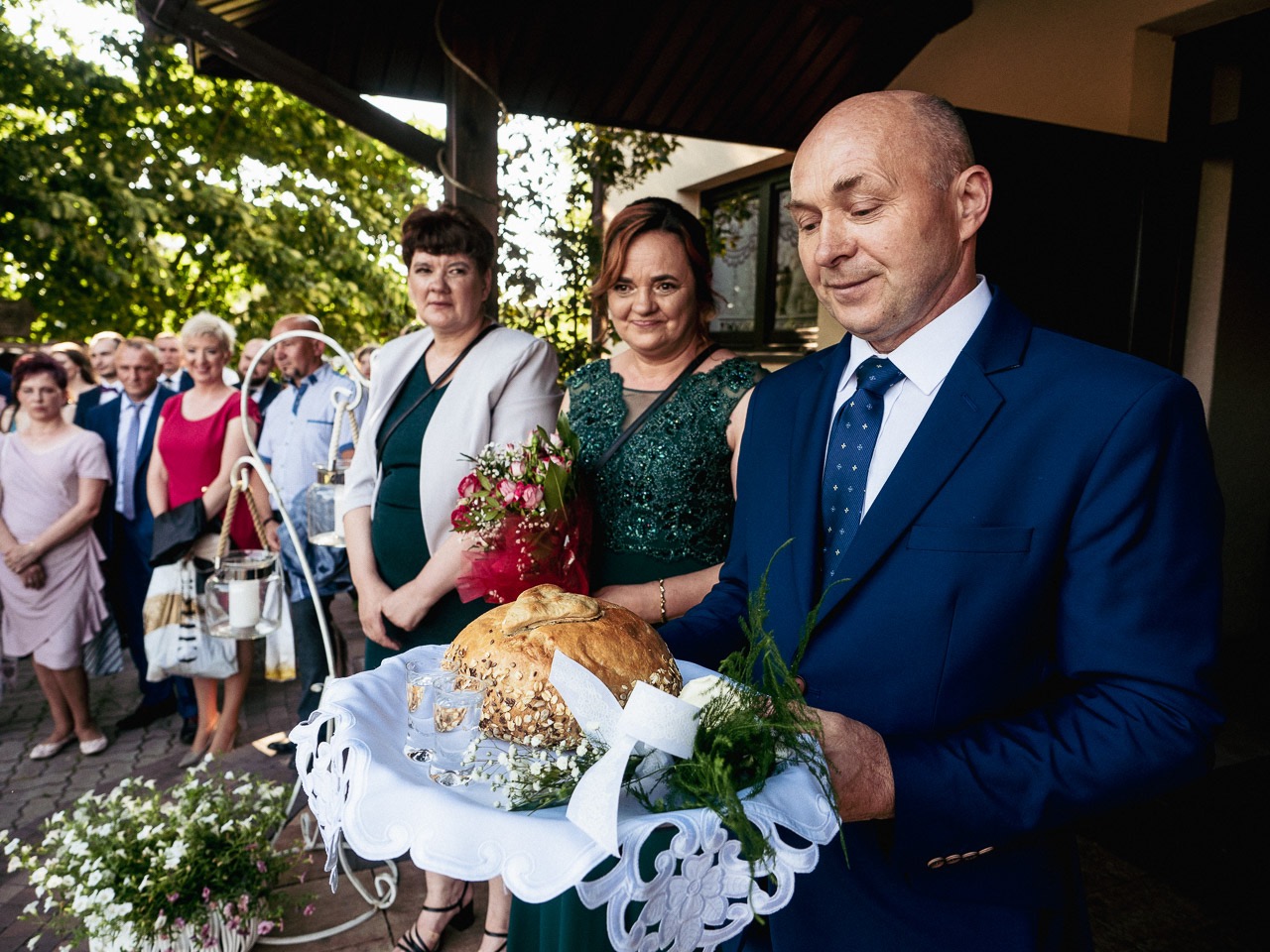 Mężczyzna w granatowym garniturze trzyma chleb weselny na tacy, za nim dwie kobiety w eleganckich sukienkach i goście weselni w tle.