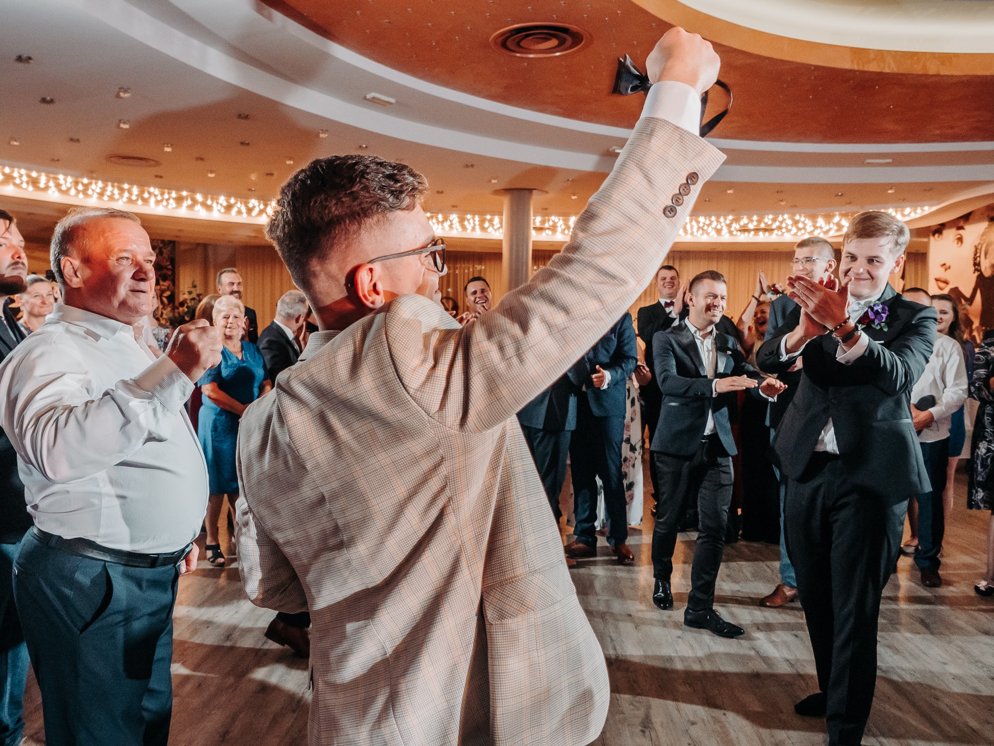Mężczyzna w kraciastej marynarce unosi krawat podczas weselnego przyjęcia, w tle goście weselni klaszczą i śmieją się.
