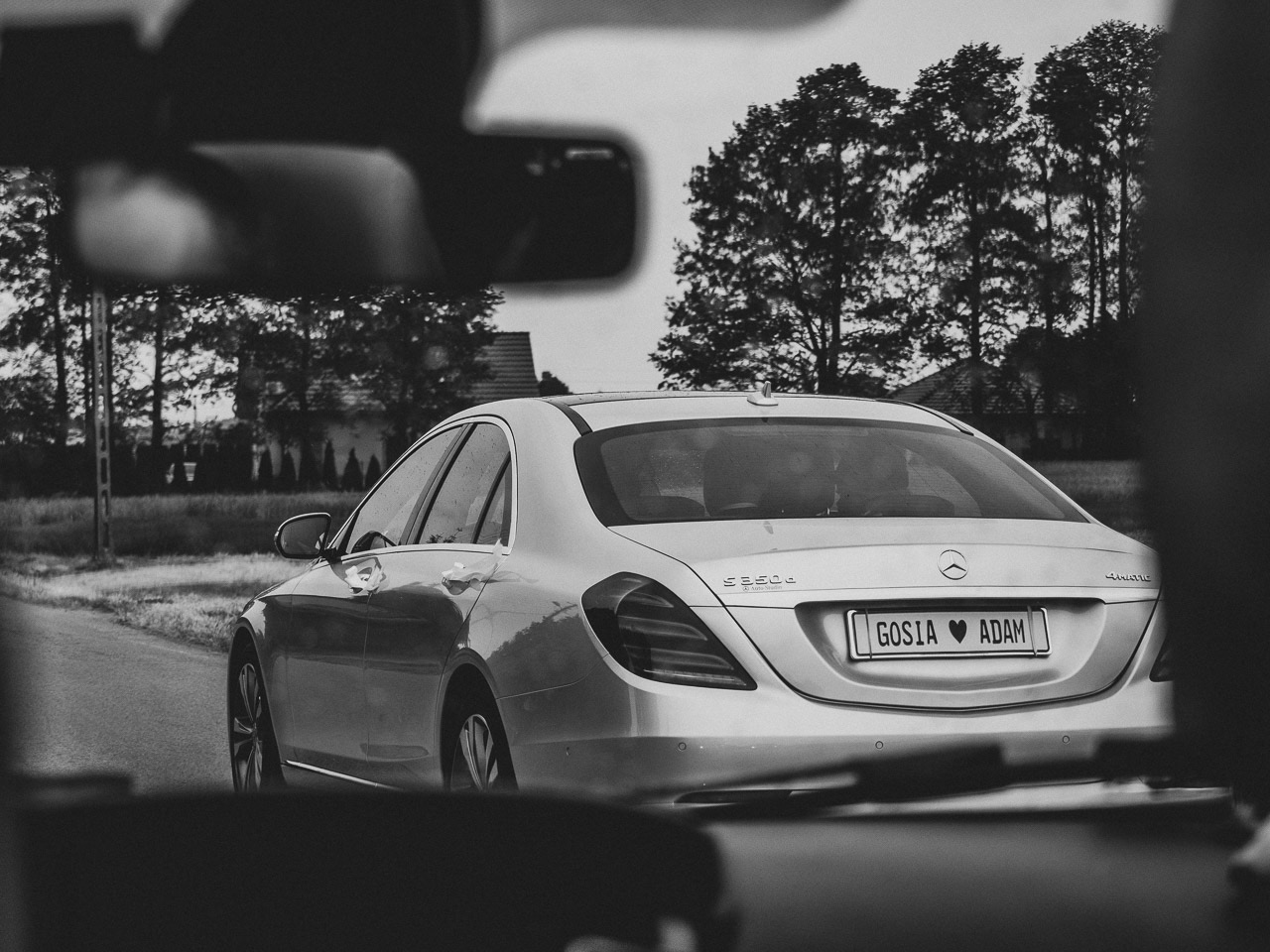 Srebrny Mercedes S350d z tablicą rejestracyjną 'GOSIA ❤ ADAM' odjeżdża po ceremonii ślubnej, widziany z wnętrza innego samochodu, w tle drzewa i budynek.