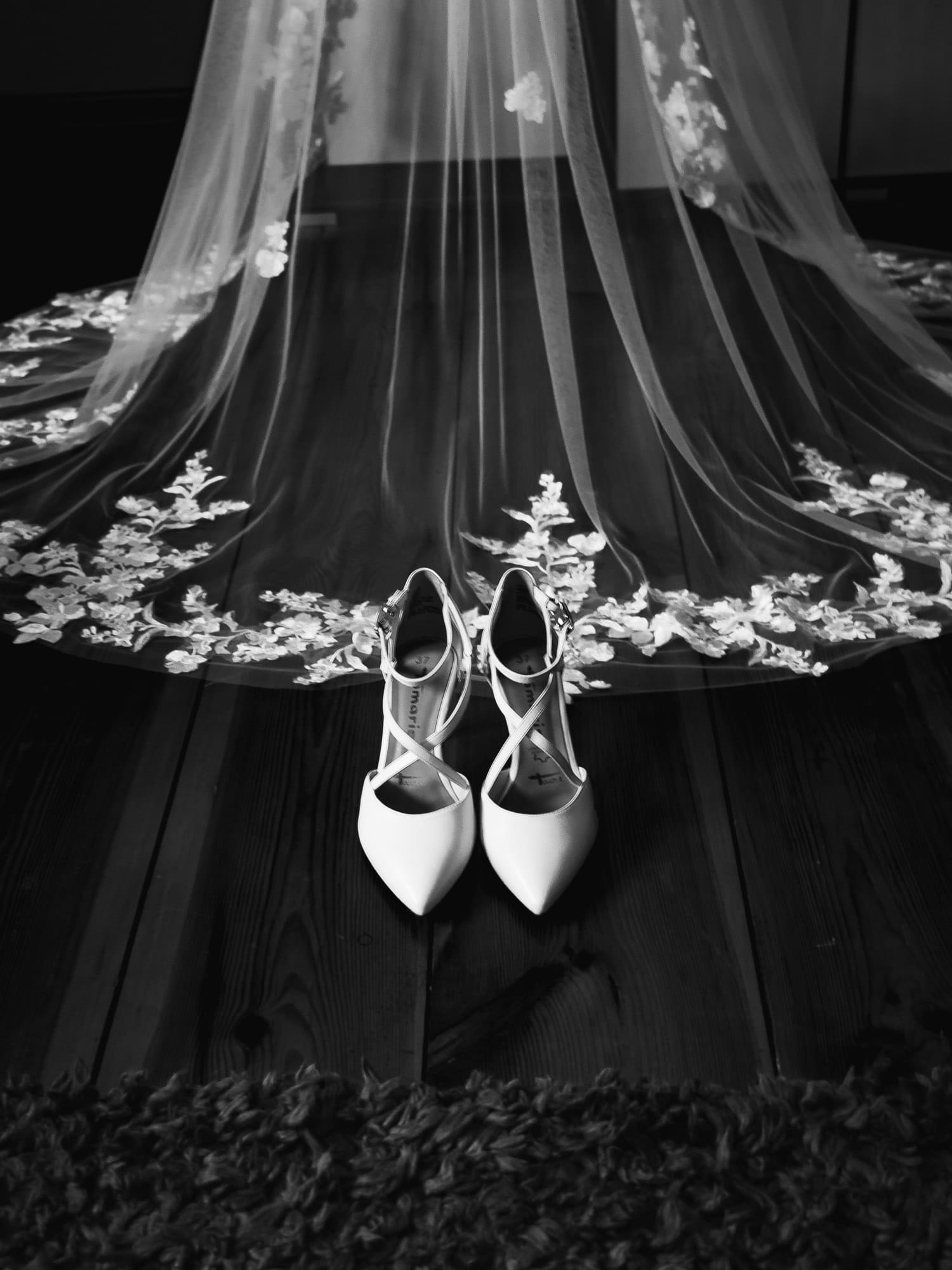 Eleganckie białe szpilki z paskami na tle drewnianej podłogi i fragmentu welonu ślubnego z koronkowymi zdobieniami w czarno-białej tonacji.