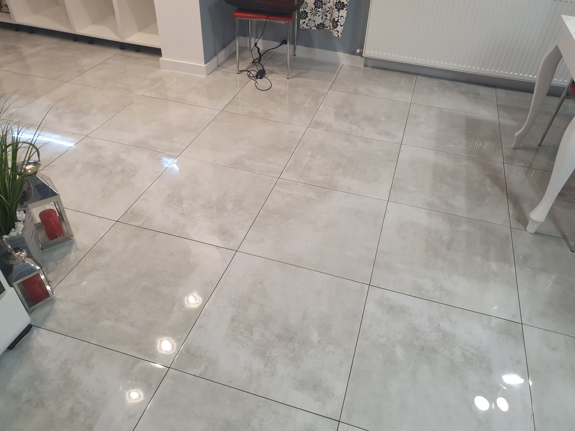 Jasnoszare, matowe płytki podłogowe ułożone w kwadratową siatkę, z delikatnym wzorem imitującym kamień, widoczne w salonie z elementami dekoracyjnymi i meblami w tle.