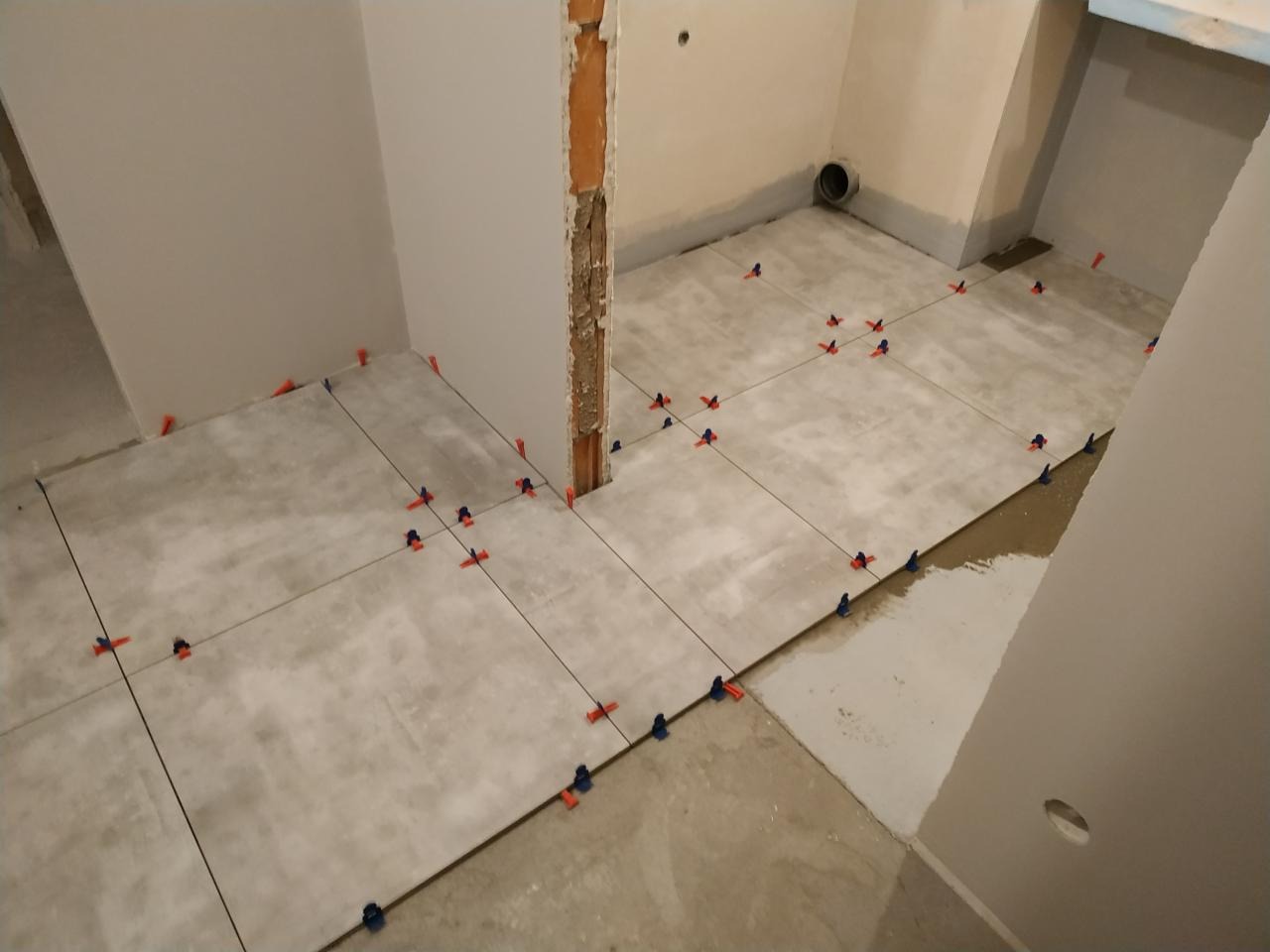Układanie szarych płytek podłogowych z użyciem krzyżyków dystansowych w trakcie remontu. Widoczne niedokończone ściany i surowa podłoga.