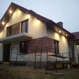 Dwukondygnacyjny dom jednorodzinny z oświetleniem zewnętrznym i fragmentami elewacji wykończonymi kamieniem dekoracyjnym, w trakcie prac wykończeniowych, widoczne rusztowania.