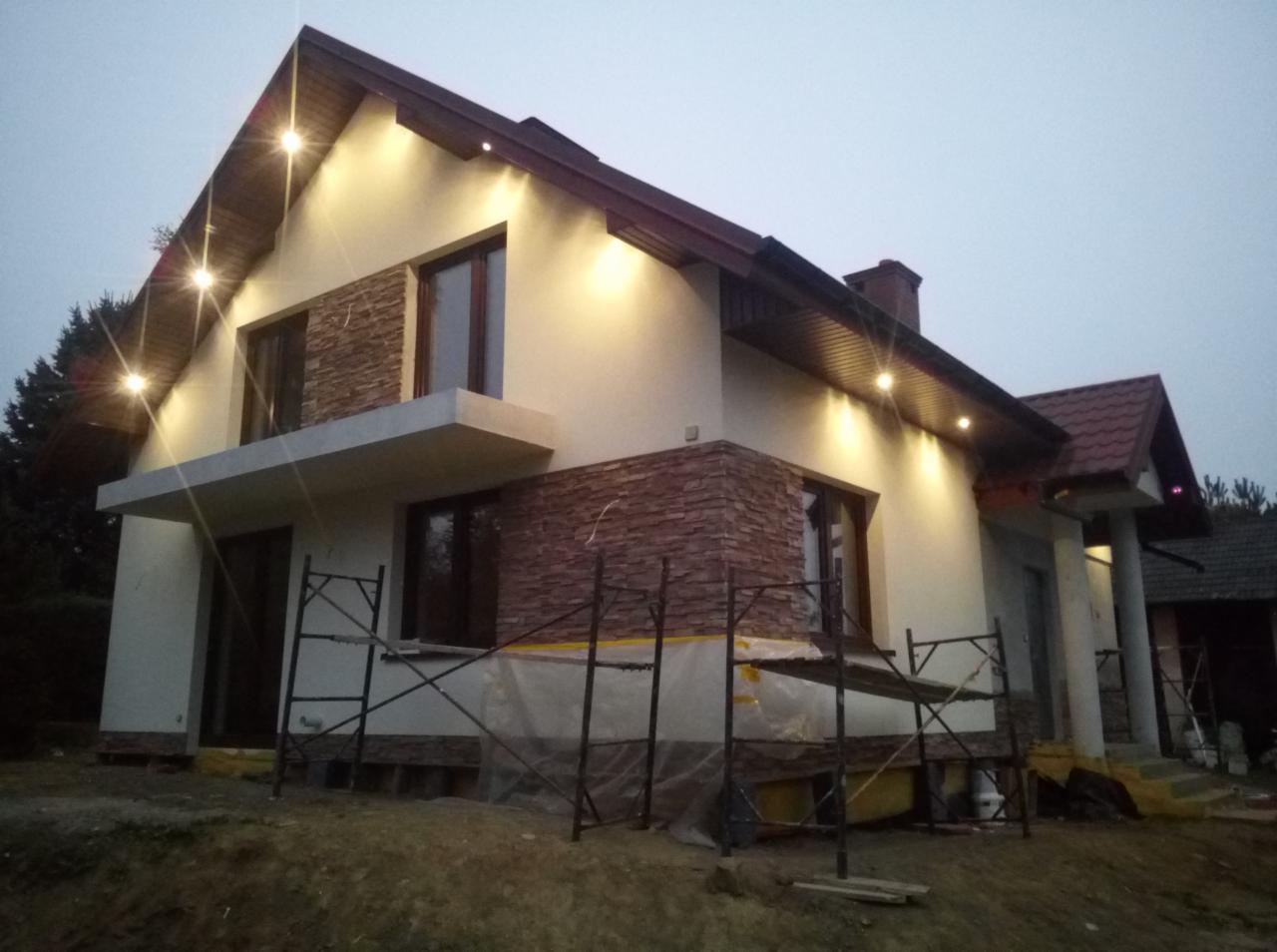 Dwukondygnacyjny dom jednorodzinny z oświetleniem zewnętrznym i fragmentami elewacji wykończonymi kamieniem dekoracyjnym, w trakcie prac wykończeniowych, widoczne rusztowania.