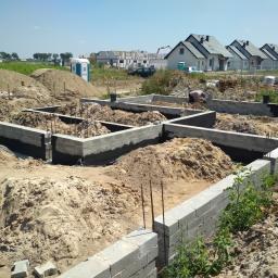 Korta - Fundamenty domu jednorodzinnego w trakcie budowy, widoczne bloczki betonowe, wykopane rowy zasypane piaskiem, w tle budynki w trakcie budowy i TOI TOI.