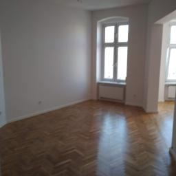 Korta - Pusty pokój z parkietem ułożonym w jodełkę, świeżo pomalowane ściany na biało, okno z białymi ramami i grzejnikiem pod nim, widok na kolejne pomieszczenie z oknem.