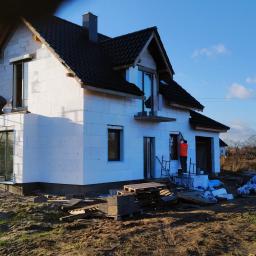 Korta - Budowa domu jednorodzinnego z białych bloczków, ocieplanie ścian zewnętrznych styropianem, pracownicy na rusztowaniach i drabinach, błękitne niebo w tle.