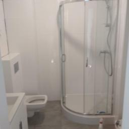 Korta - Nowoczesna, wykafelkowana łazienka z kabiną prysznicową z zaokrąglonymi szybami, umywalką z szafką i podwieszaną toaletą. Na podłodze widoczne środki czystości.