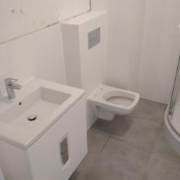 Korta - Nowoczesna, minimalistyczna łazienka z białą armaturą: umywalka z szafką, podwieszana toaleta z przyciskiem spłukującym i fragment kabiny prysznicowej, na tle białych płytek ściennych i szarej...