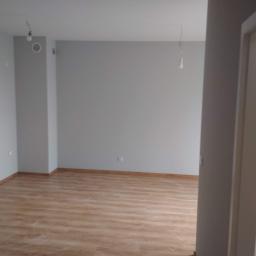 Korta - Pomalowane na szaro puste pomieszczenie z ułożonymi panelami podłogowymi, widoczne wystające kable od oświetlenia sufitowego i otwarte białe drzwi.