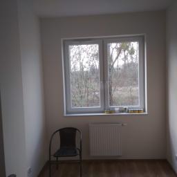 Korta - Pomalowane na biało wnętrze pokoju z brązową podłogą, szarym krzesłem i białym grzejnikiem pod oknem, na parapecie stoją puszki z farbą.