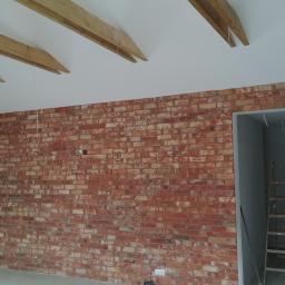 Korta - Ściana z czerwonej cegły dekoracyjnej w surowym wnętrzu z drewnianymi belkami sufitowymi i drabiną w przejściu do kolejnego pomieszczenia.