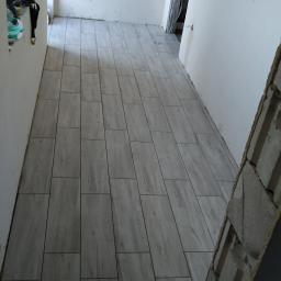 Korta - Korytarz w trakcie wykańczania z ułożoną podłogą z płytek imitujących drewno, widoczne wystające kable i ściana z pustaków.