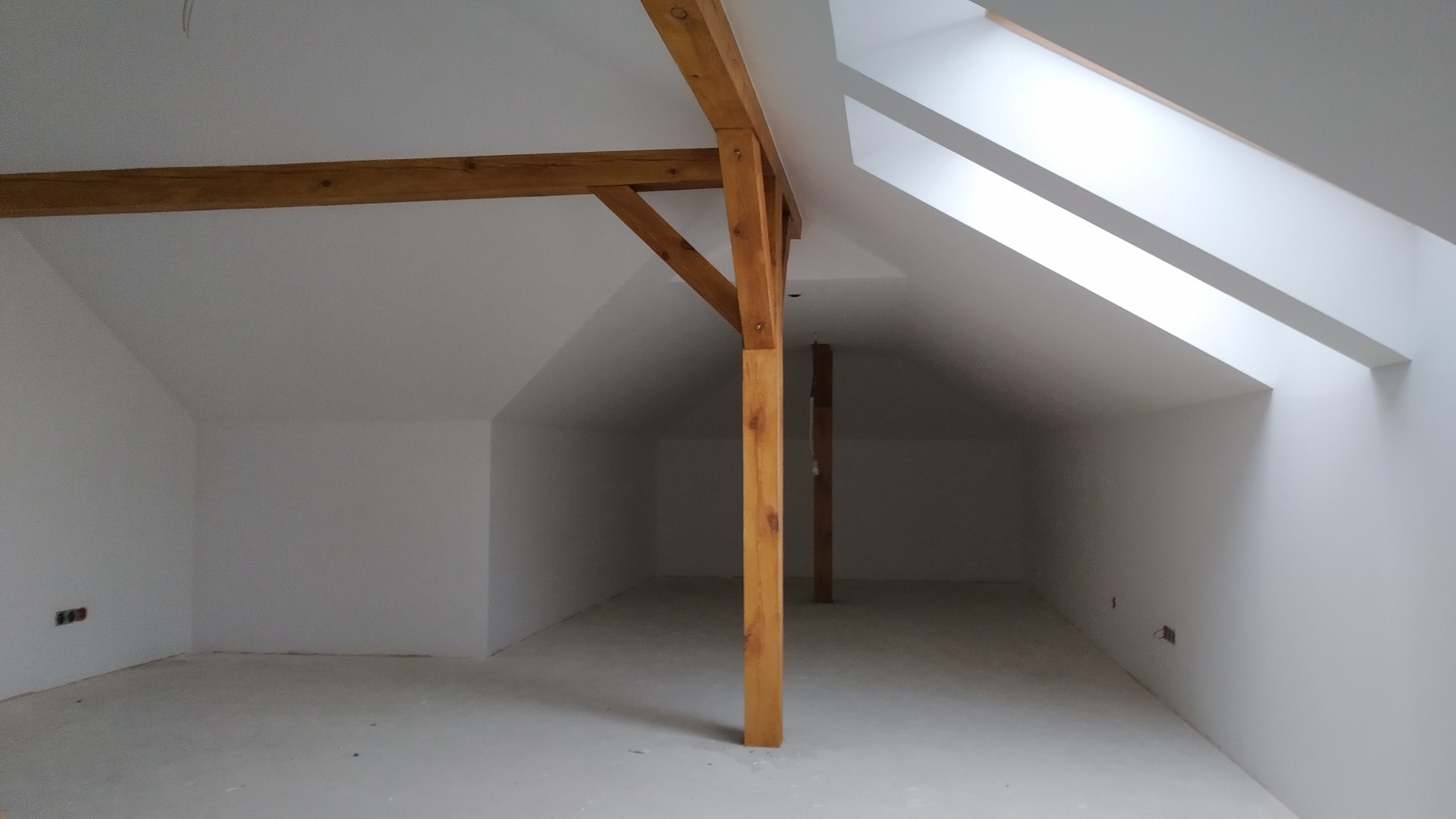 Poddasze po remoncie, białe ściany i sufit, drewniane belki konstrukcyjne, okno dachowe w skosie dachu, widoczne gniazdka elektryczne w ścianie, surowa podłoga.