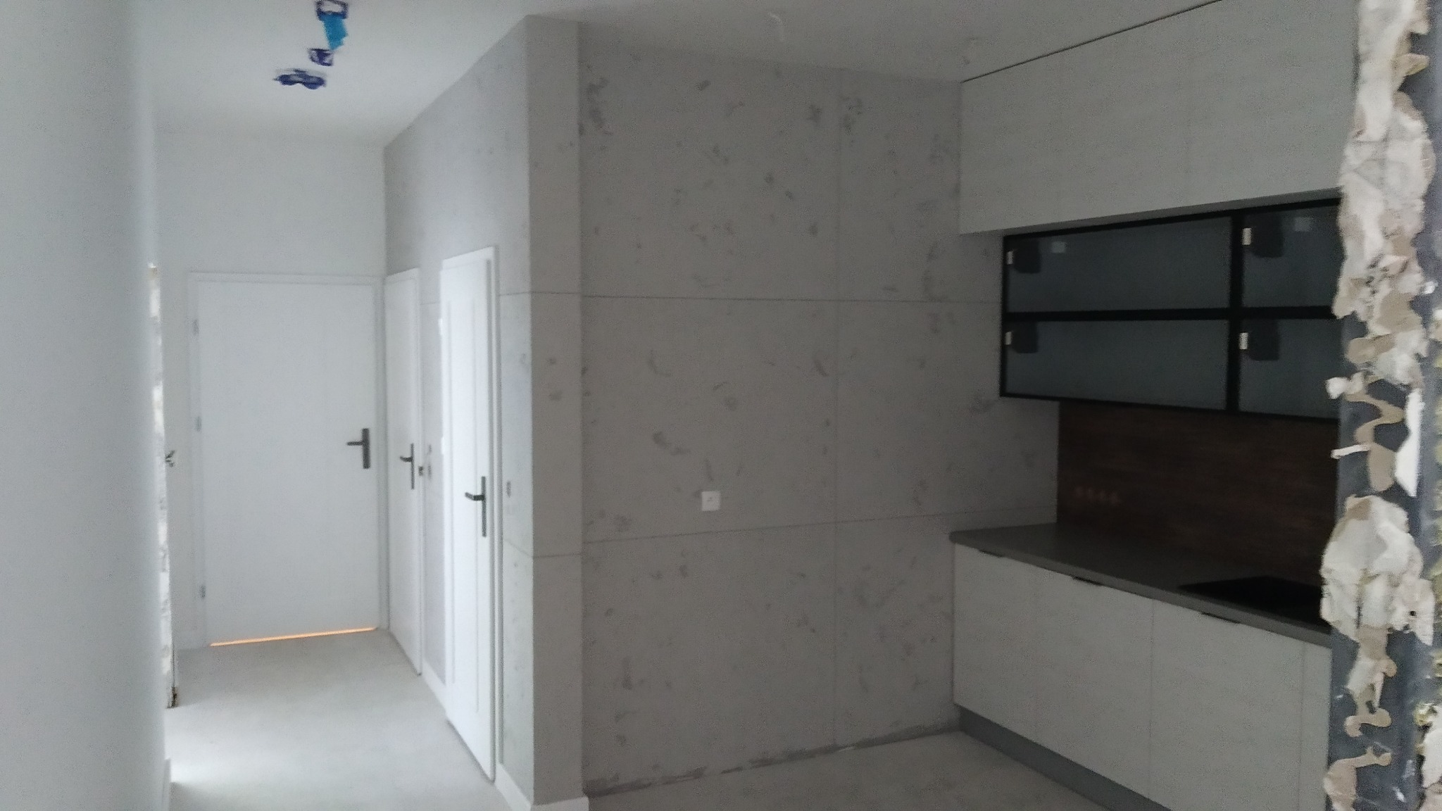 Wnętrze w trakcie wykończenia: widoczna zabudowa kuchenna z szarymi frontami i czarnymi ramami szafek, ściany pokryte szarą, wzorzystą okładziną, obok białe drzwi, fragment ściany z resztkami...