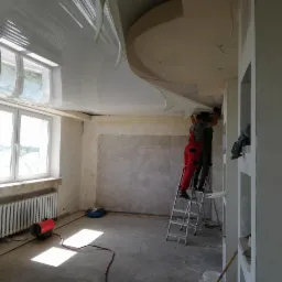 Montaż błyszczącego sufitu napinanego w jasnym pomieszczeniu, dwóch monterów na drabinie, widoczny grzejnik i nagrzewnica.