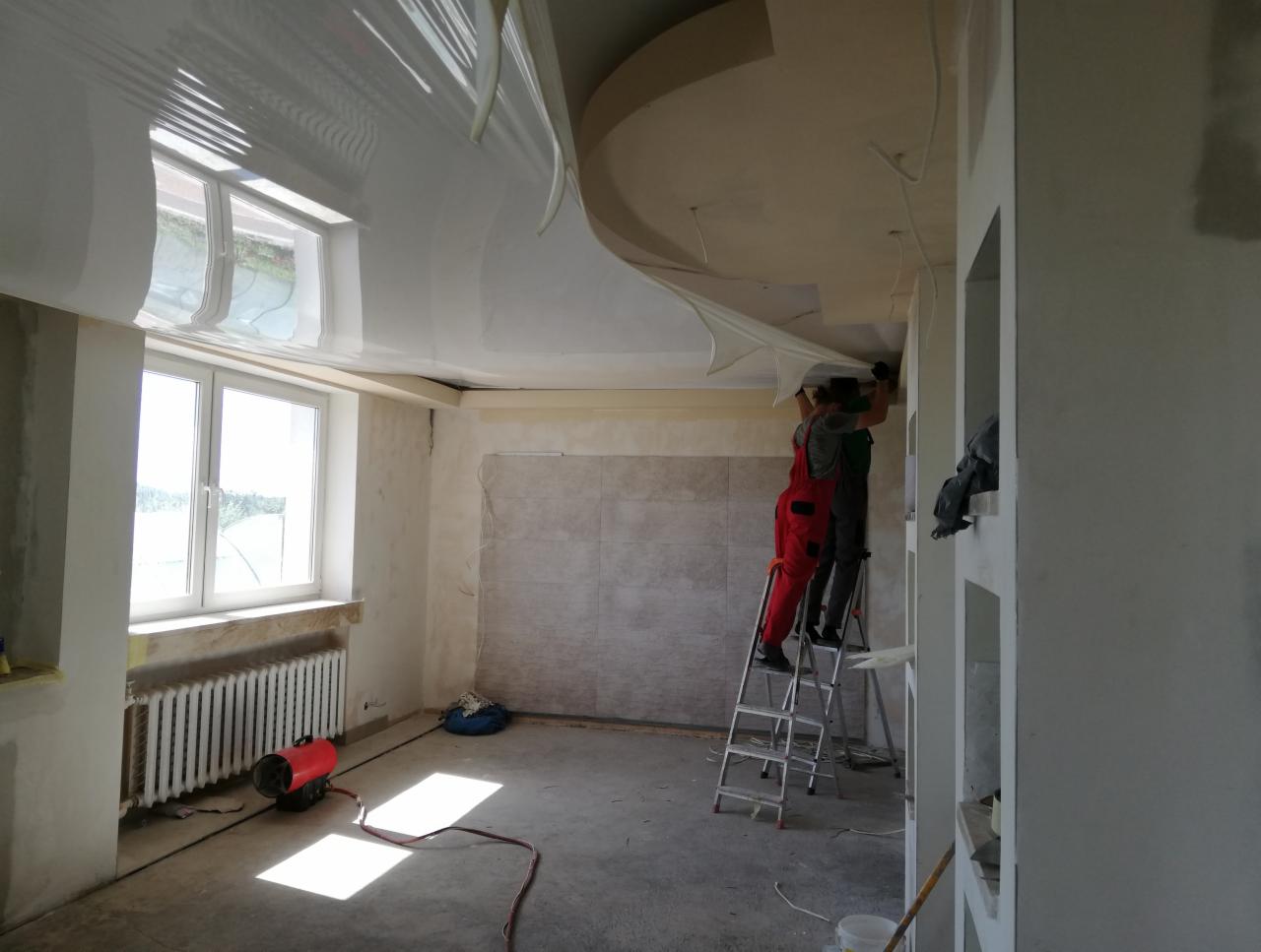 Montaż błyszczącego sufitu napinanego w jasnym pomieszczeniu, dwóch monterów na drabinie, widoczny grzejnik i nagrzewnica.