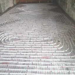 Rozłożone białe rury ogrzewania podłogowego na folii z czerwoną kratką, mocowane czarnymi klipsami do podłoża, widoczne surowe ściany w tle.