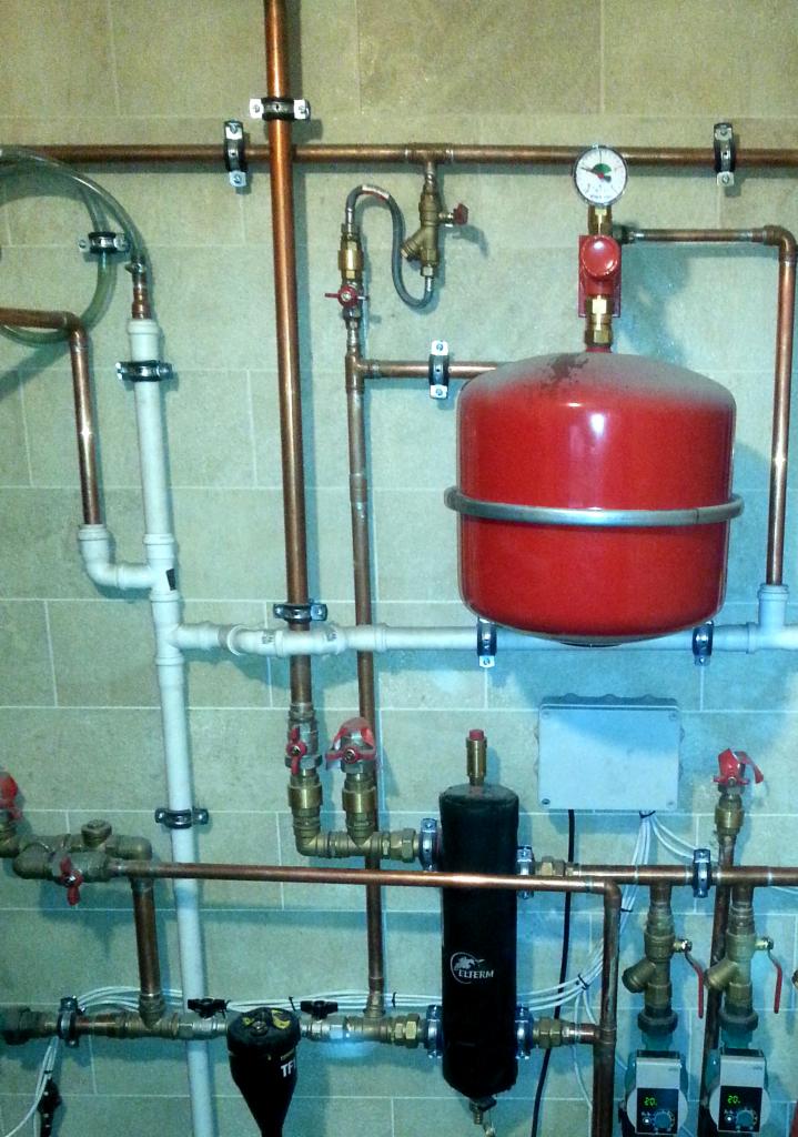 Kompleksowy system rur miedzianych i plastikowych, z czerwonym zbiornikiem ciśnieniowym i czarnym filtrem, zamontowany na ścianie z płytek ceramicznych.