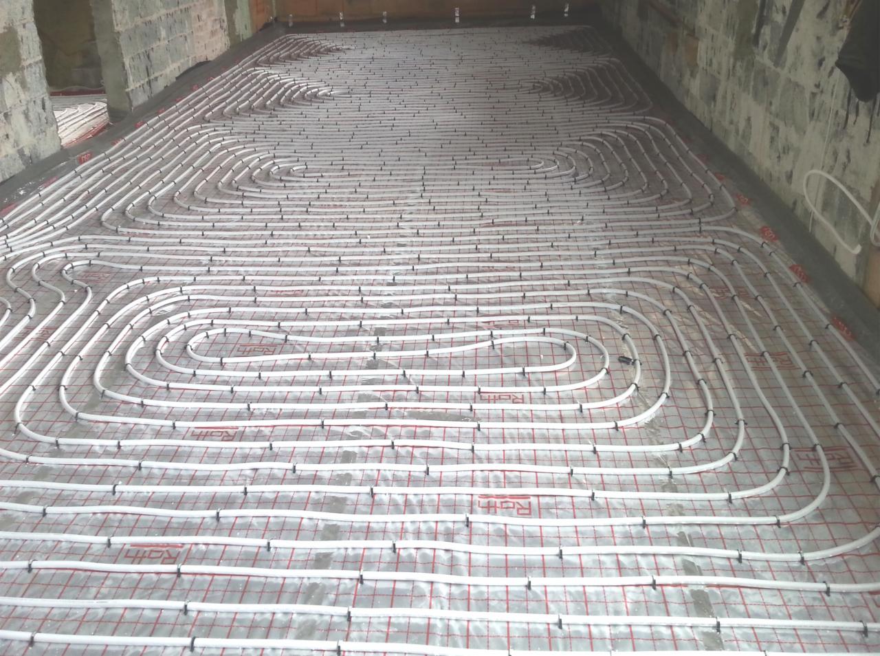 Rozłożone białe rury ogrzewania podłogowego na folii z czerwoną kratką, mocowane czarnymi klipsami do podłoża, widoczne surowe ściany w tle.