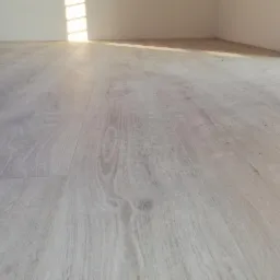 Jasne, drewnopodobne panele podłogowe ułożone w nowym pomieszczeniu, widoczne gniazdka elektryczne na białej ścianie i delikatne zabrudzenia na powierzchni.