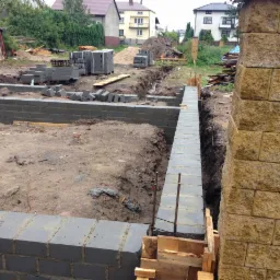 Wykopane fundamenty z częściowo wylanych ścianami z betonowych bloczków, widoczne zbrojenie i szalunki, tło stanowią domy i drzewa.