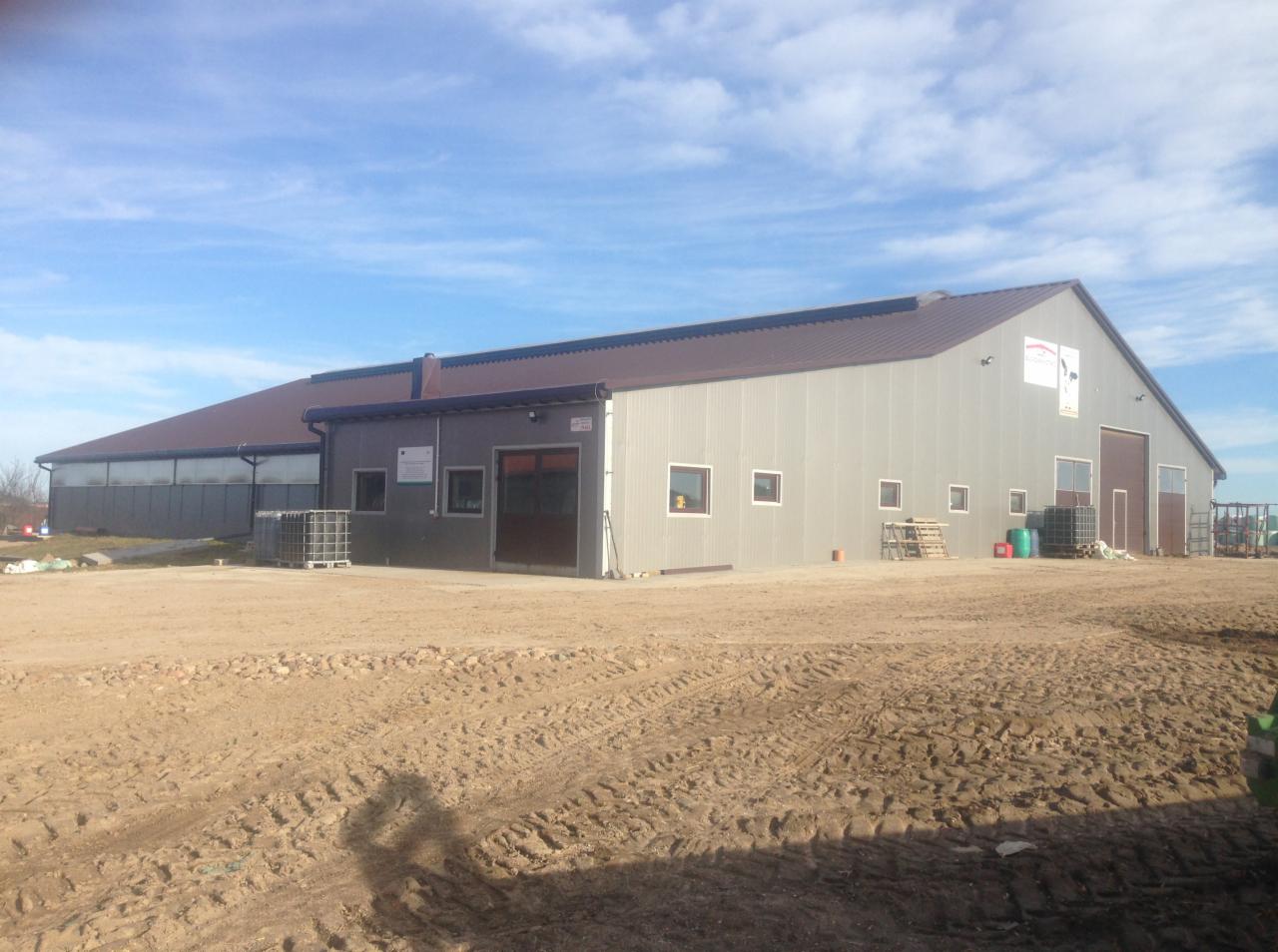 Duży, szary budynek magazynowy z brązowym dachem i bramami, widoczny z zewnątrz na placu budowy z ziemią i śladami opon.