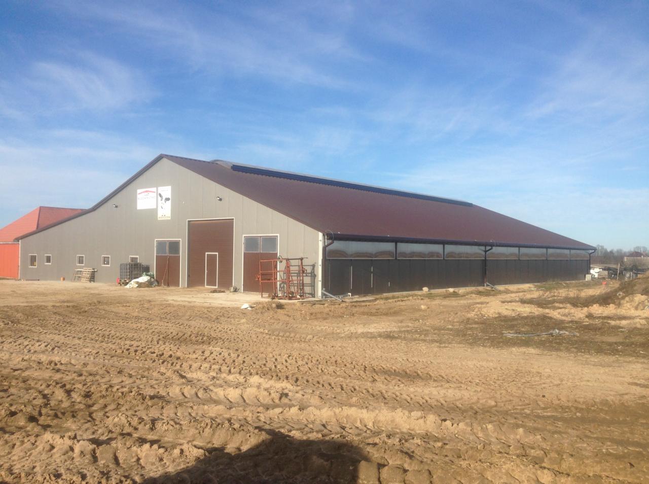 Duży budynek gospodarczy z szarymi ścianami i brązowym dachem, widoczny logo firmy na ścianie, teren przed budynkiem częściowo utwardzony