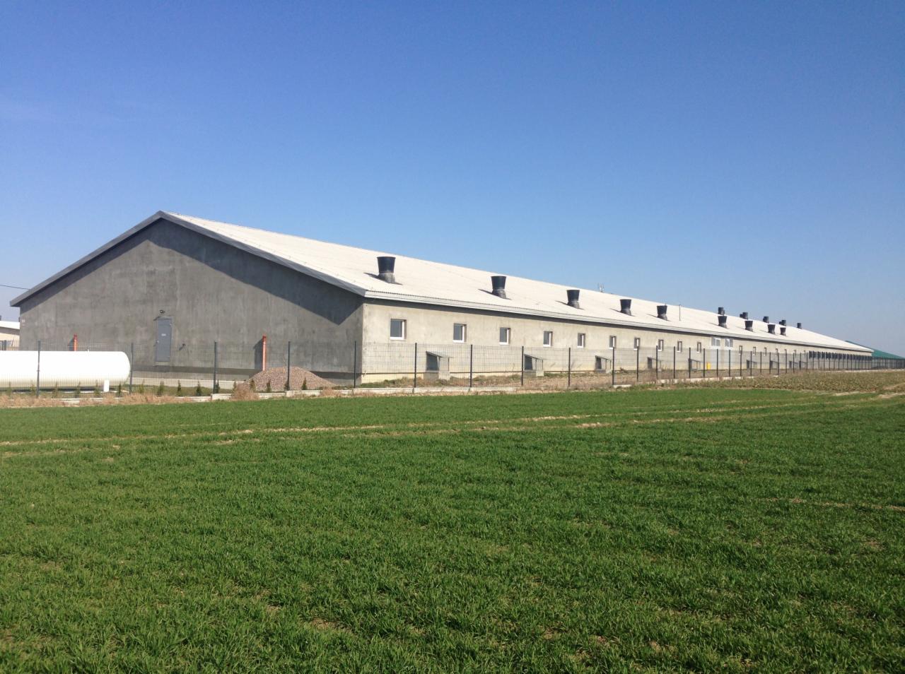 Długi, szary budynek przemysłowy z białym dachem i kominami, otoczony zielonym polem i ogrodzeniem z siatki.