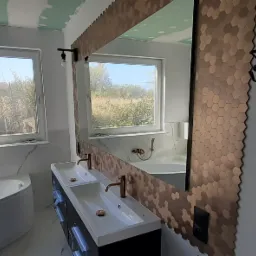 Łazienka w trakcie wykańczania: widoczna wanna, umywalka z dwoma kranami w kolorze miedzi, lustro z ozdobną ramą z heksagonów, surowy sufit i okno z widokiem na zewnątrz.