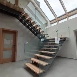 Nowoczesne schody z drewnianymi stopniami i szklanymi balustradami, oświetlone naturalnym światłem z okien dachowych, widok z dołu.