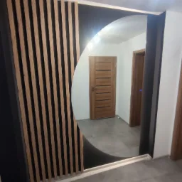 Nowoczesna aranżacja przedpokoju z lustrem o nietypowym, półokrągłym kształcie, obudowanym czarną ramą i drewnianymi lamelami, widoczne drzwi wewnętrzne.