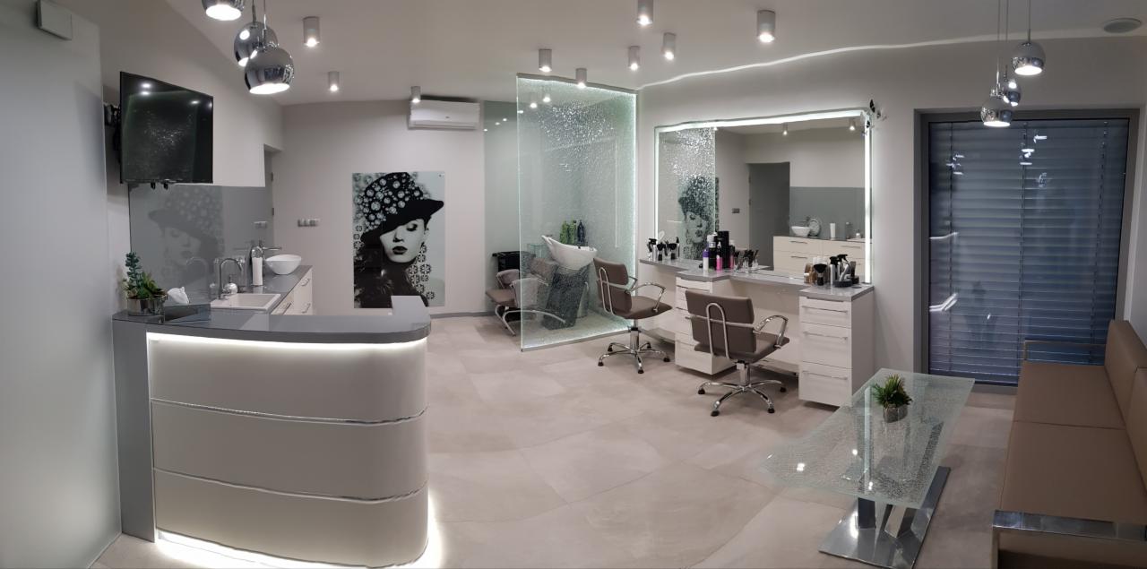 Nowoczesne wnętrze salonu fryzjerskiego z recepcją z podświetleniem LED, szklaną ścianką działową, stanowiskami z lustrami i fotelami fryzjerskimi oraz szklanym stolikiem kawowym.