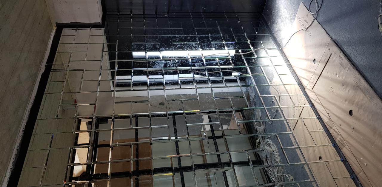 Montaż mozaiki lustrzanej z kwadratowych płytek na ścianie, widoczne kable i narzędzia montażowe, w trakcie prac wykończeniowych.