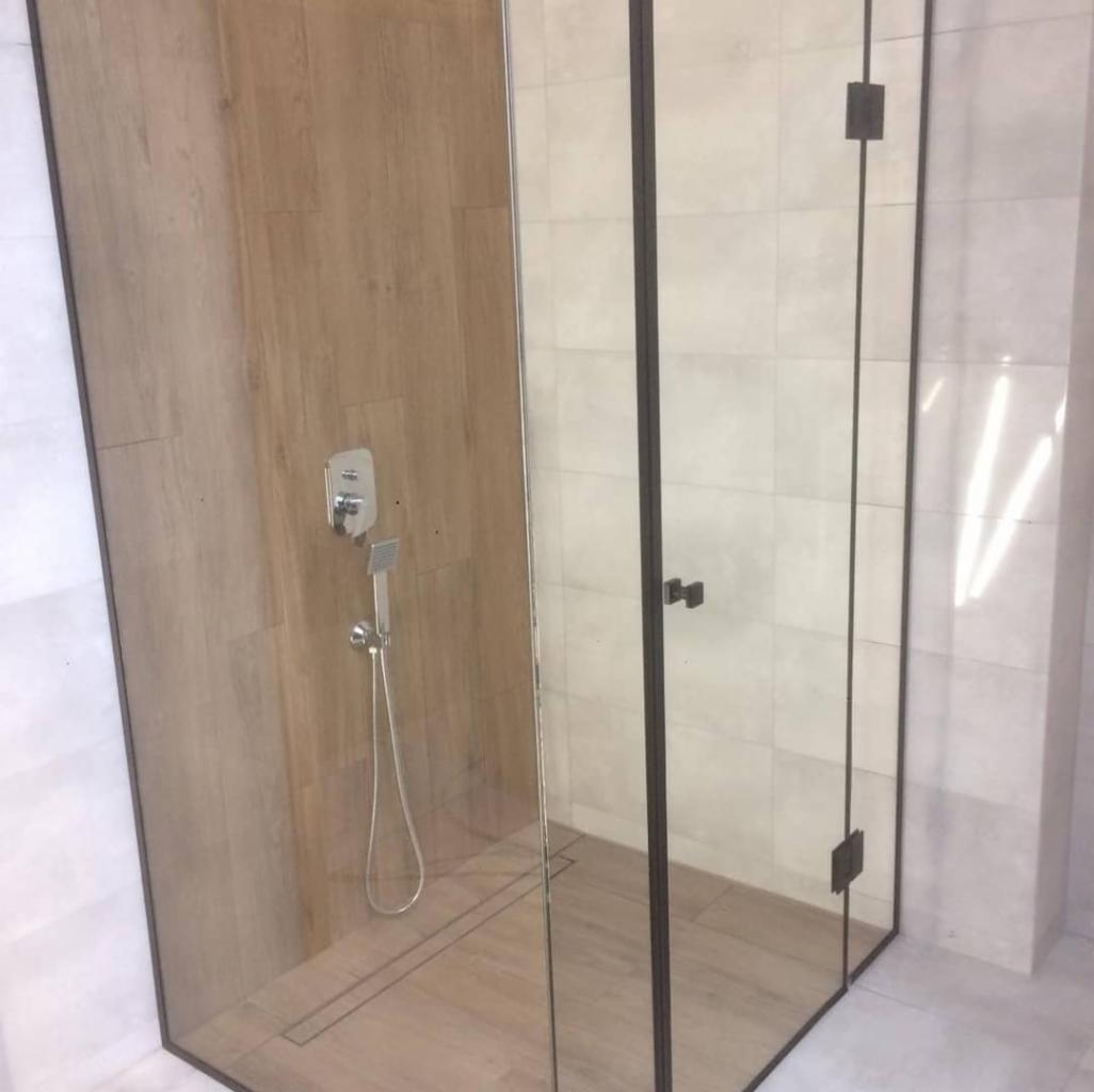 Kabina prysznicowa z czarnymi profilami, wykończona płytkami imitującymi drewno i białymi płytkami, z odpływem liniowym w podłodze.