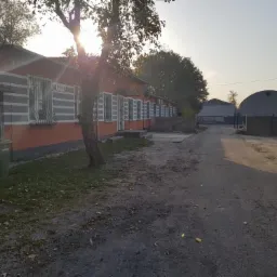 Widok na budynek z pomarańczową elewacją, szare pasy pod oknami i metalowe kraty w oknach, z widoczną drogą dojazdową, samochodem po prawej i konstrukcjami namiotowymi w tle, w słoneczny dzień.